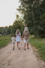 Fototapeta premium A family walks along a dirt path in a rural area. The group includes a blond woman, a young boy, and a man carrying a child on his shoulders.