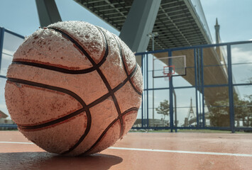 A heavily used basketball rests on an outdoor court, with a hoop and fiberglass backboard visible beneath the industrial Ring Road Bridge in the background. Space for text.