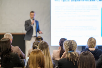 Business presentation conference room. Speaker with microphone addressing audience. Large projected slide on screen conveys information to attendees.