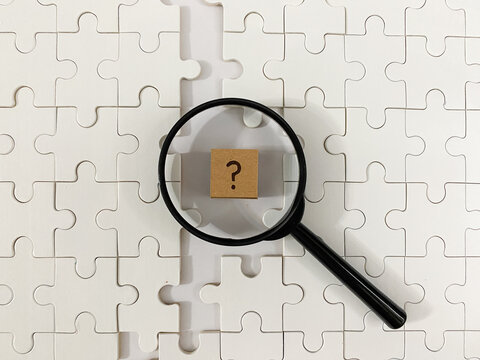 Magnifying glass focuses on a cardboard question mark block atop white jigsaw pieces, representing business investigation, problem-solving, strategic research, and finding answers complex projects 