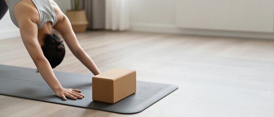 Young adult woman practicing downward facing dog yoga pose on a grey mat with a cork block in a bright minimalist living room during International Yoga Day.