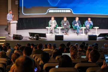 Panel discussion on stage featuring five speakers in green chairs, audience in auditorium watches presentation, blue screen behind stage