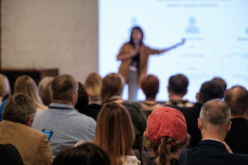 Presenter speaks to large audience during conference. Attendees listen, take notes. Stage...