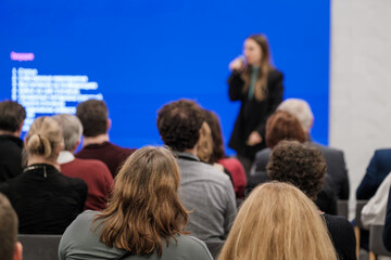 Audience focuses on speaker during conference against bright blue screen. Attendees sit in modern indoor setting paying attention to presentation.