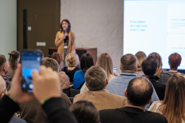 A speaker presents to a crowded conference room while attendees watch large screens and capture moments with mobile devices. The scene conveys learning, engagement, and professional gathering.