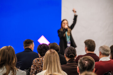Presenter speaks to large audience on stage with bright blue backdrop. Attentive listeners form rows in conference setting. Energetic mood and clear communication drive the session.