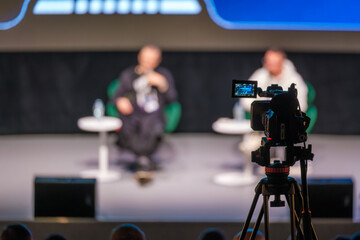 Panel discussion on stage captured by camera on tripod while speakers talk and audience watches