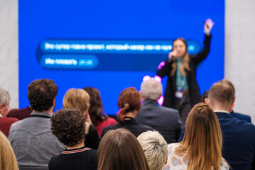 Speaker on stage addresses crowd at conference while audience listens. Bright blue backdrop highlights presentation. Energetic atmosphere reflects learning and networking.