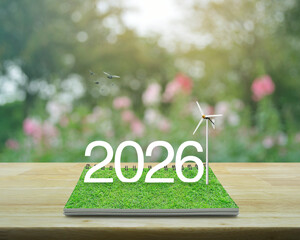 2026 white text with wind turbine on green grass on open book on wooden table over blur pink flower and tree in garden, Happy new year 2026 ecology saving power and energy concept