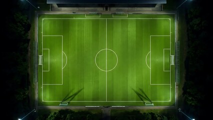 Vibrant green football pitch illuminated at night, captured from an overhead drone perspective, showcasing the field lines and bright stadium lights ready for a game