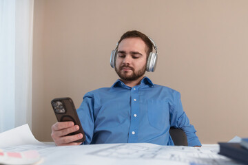 Young man in a blue shirt relaxing at a desk, using a smartphone and wearing headphones indoors.