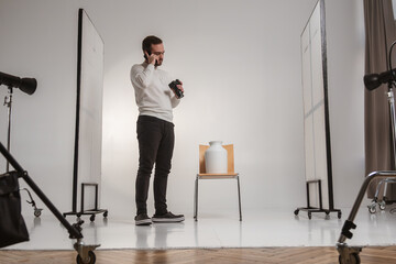 A photographer stands in a studio with equipment, holding a camera and phone, preparing for a product shoot.