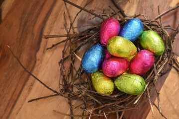 Foil wrapped colourful easter eggs in pink, green, blue and yelow in a natural nest made of sticks and twigs, against a multi grain brown wooden background.