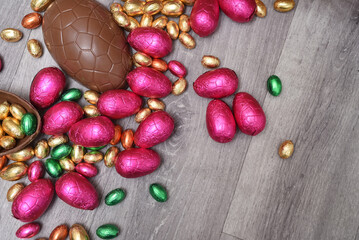 Selection of colourful foil wrapped chocolate easter eggs in pink, red, gold and green. Large halves of a brown milk chocolate egg have mini eggs inside, on a grey wooden background.