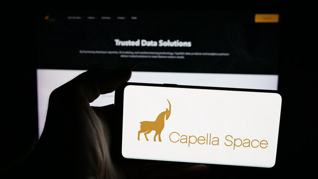 Stuttgart, Germany - 12-17-2025: Person holding mobile phone with logo of US satellite company Capella Space on screen in front of web page. Focus on phone display.