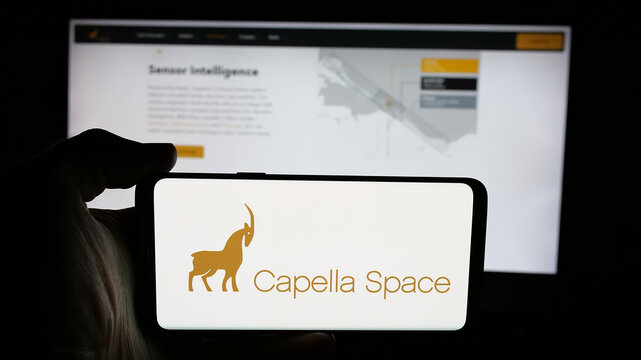 Stuttgart, Germany - 12-17-2025: Person holding cellphone with logo of US satellite company Capella Space on screen in front of webpage. Focus on phone display.