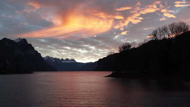 Walensee Switzerland lake at sunset with orange clouds reflecting on calm water, mountain silhouettes, peaceful evening light, serene European landscape nature view.