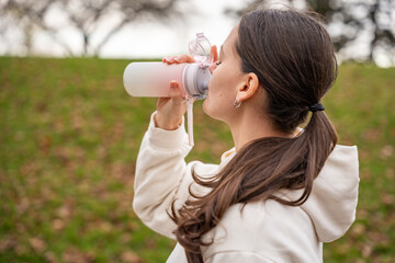 Woman drinking water after jogging in park. Hydrating after cardio session in park. Essential healthy habit for maintaining body balance.
