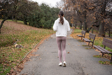 Fototapeta premium Woman jogging in autumn park wearing white hoodie. Running along path in peaceful city park, showcasing active lifestyle and self care outdoors.