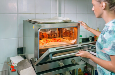 Unrecognizable female chef opening professional oven door to monitoring freshly baked pizza in commercial kitchen, expertly preparing a delicious meal for eager customers