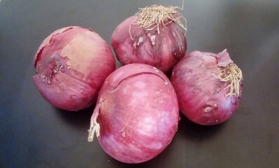 Four red onions with dry skin and roots on a dark surface with subtle shadows and a neutral mood.