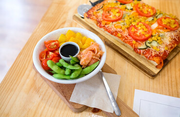 Top view of poke bowl with fresh salmon, mango, edamame, cherry tomatoes and soy sauce, serving on restaurant table next to vegetarian focaccia, in casual lunch. Organic and natural fast food concept