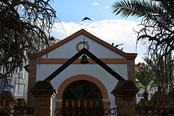 Pigeons flying over a shrine in Spain with a cross and a bell

