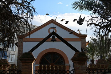 Pigeons flying over a shrine in Spain with a cross and a bell

