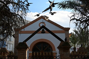Pigeons flying over a shrine in Spain with a cross and a bell

