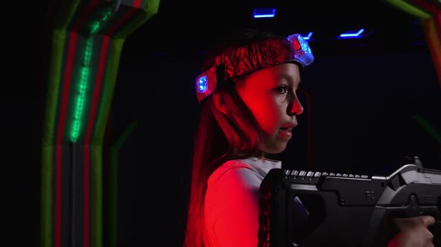 An active girl is playing laser tag with friends, she holds a laser weapon and takes aim moving in a circle. A playground with bright stripes on the wall. A playground for active games for children.