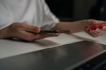 Close-up cropped shot of person holding smartphone in one hand and red credit card in other while preparing online purchase on laptop, illustrating cashless e-commerce and digital payment convenience.