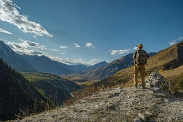 A lone traveler in hiking gear stands at a mountain road's edge, contemplating a vast autumn valley. Rear-side view captures the figure overlooking forests, rivers, and peaks.