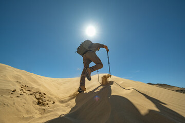 A hiker with poles and a backpack ascends a sand dune under a bright sun in a cloudless blue sky. Low-angle general shot from behind captures the rising sand with each step.