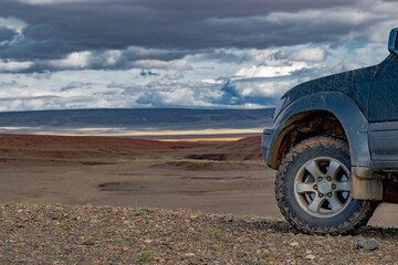 An off-road vehicle is parked on a dirt rise overlooking the endless Mongolian steppe. A medium shot of its left wheel clad in a mud tire stands against dark stormy clouds.