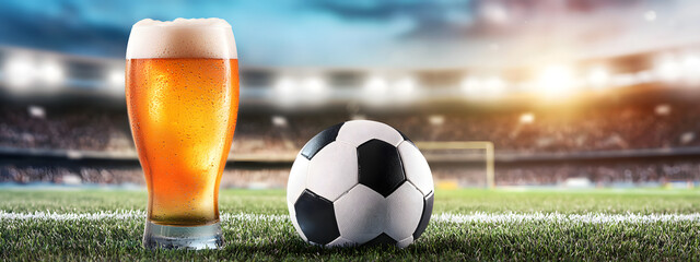 A glass of beer next to a football ball on the grass of a football field against the backdrop of stands and floodlights with a sunny sky - a creative football banner