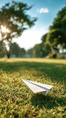 A serene scene featuring a paper airplane resting on lush green grass, surrounded by trees under a bright blue sky.