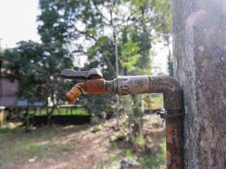 ​Vintage rusty water tap on a concrete post in a lush garden. Old outdoor metal faucet with weathered plumbing pipe in a rural backyard setting