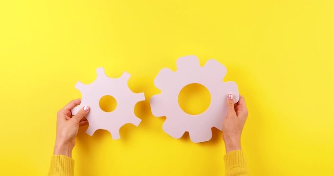 Close-up of female hands holding a gear symbol on a bright yellow background. Conceptual 4K video representing setup, repair, maintenance, engineering, and problem-solving processes.