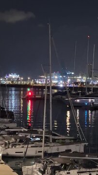 Night view of Barcelona harbor with marina boats and industrial port lights reflecting on calm water, urban waterfront scene after dark showing city infrastructure, maritime activity and Mediterranean