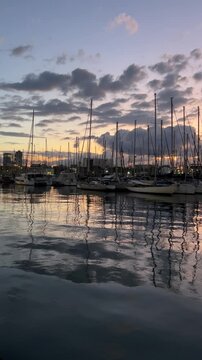 Sunset marina with sailboats and yachts reflected on calm water under dramatic cloudy sky, peaceful harbor landscape at dusk with moored boats, nautical atmosphere and cinematic travel background
