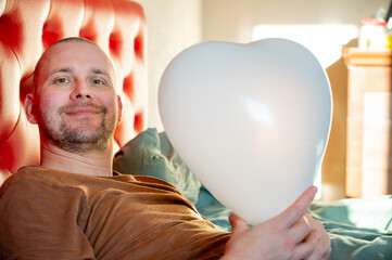 Man inflating heart-shaped balloons in a bedroom on a red bed, preparing a romantic setting for Valentine’s Day.
