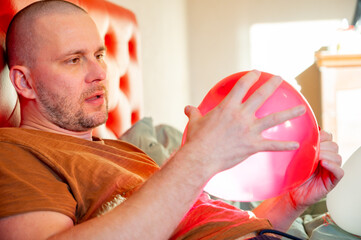 Man inflating heart-shaped balloons in a bedroom on a red bed, preparing a romantic setting for Valentine’s Day.