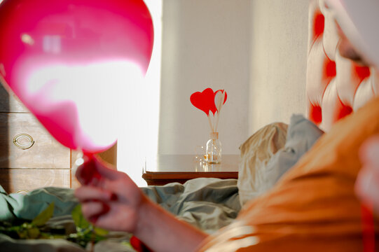 Man inflating heart-shaped balloons in a bedroom on a red bed, preparing a romantic setting for Valentine&rsquo;s Day.