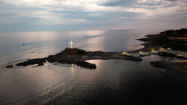 Aerial view of illuminated cross monument atop a rocky cape dividing the sea from a marina, a beacon against the dusky sky, Lozenets, Tsarevo, Bulgaria.