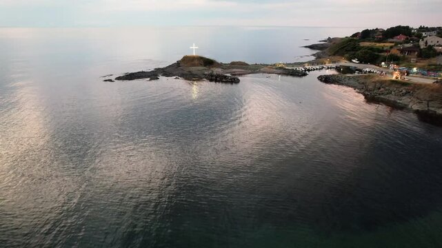 Aerial view of the white cross standing tall on a rocky peninsula surrounded by the dark, rippling sea near the harbor, Lozenets, Tsarevo, Bulgaria.