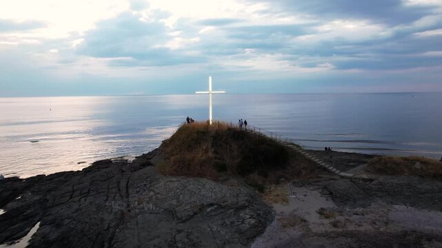 Aerial view of a glowing cross standing atop a rocky outcrop surrounded by the sea with dramatic skies, Lozenets, Tsarevo, Bulgaria.