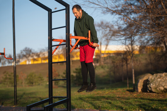 A bearded middle-aged man exercises on a sports ground in a park using exercise equipment, doing an exercise on the parallel bars - Powered by Adobe
