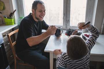 A bearded man prepares breakfast and eats at the table with his son, cheerful conversations at the table, spending time together