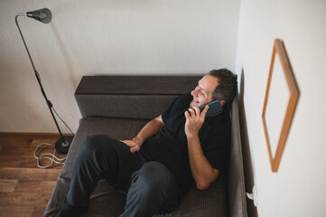 Bearded man talking on the phone at home lying on the sofa with a smile, pleasant conversation on the phone