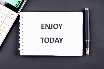 A spiral notebook displays the words ENJOY TODAY. A sleek fountain pen rests to the right of the notebook, and a calculator is partially visible on the left, all against a solid background